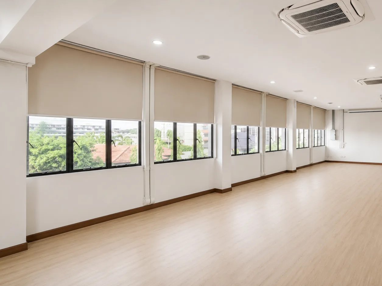 Empty open-plan office space with beige roller blinds lowered on a row of windows and light wood flooring