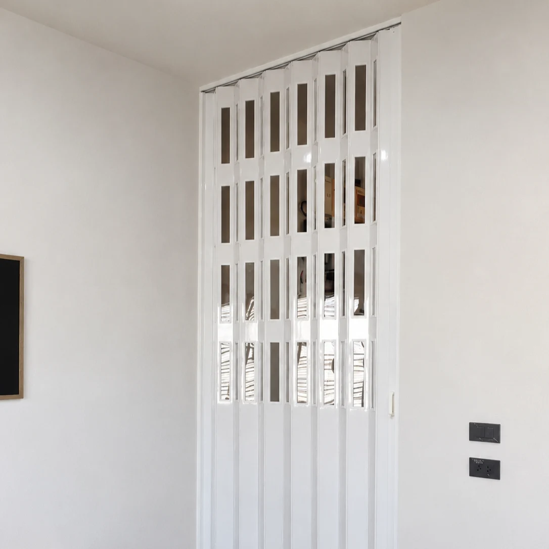 White accordion room divider with narrow vertical glass panel inserts closing a corner doorway beside a wood sideboard