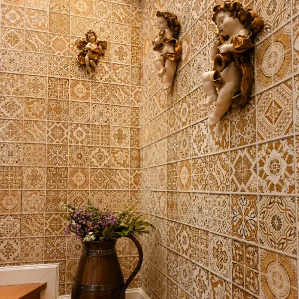 Ornate tile-print wallpaper in a stairwell corner with gilded cherub figurines mounted on the wall and a flower-filled jug
