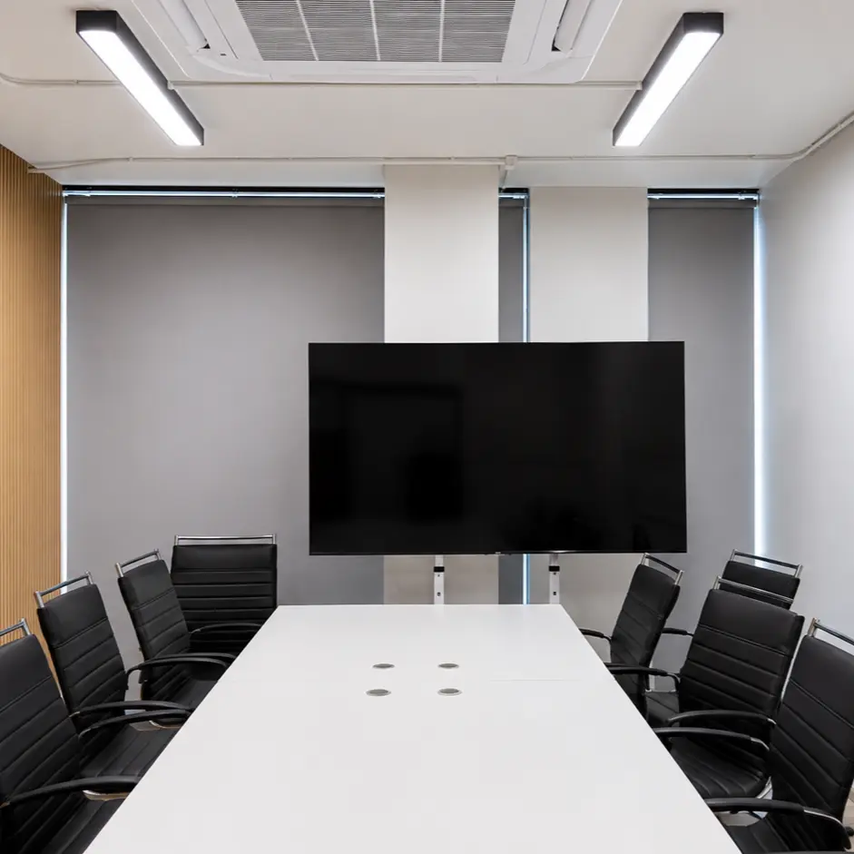 Meeting room with gray roller blinds lowered over floor-to-ceiling windows behind a wall-mounted TV and conference table, wood slat accent wall