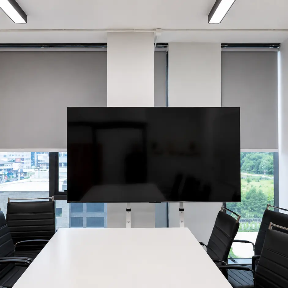 Meeting room with gray roller blinds on floor-to-ceiling windows behind a wall-mounted TV and conference table