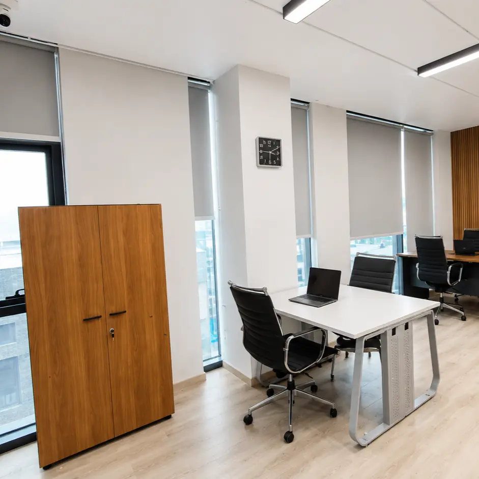Open-plan office area with gray roller blinds on floor-to-ceiling windows, workstations with laptops and a wood slat accent wall