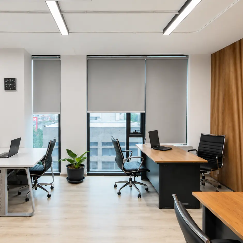 Open-plan office area with gray roller blinds lowered over floor-to-ceiling windows, workstations with laptops and a wood cabinet