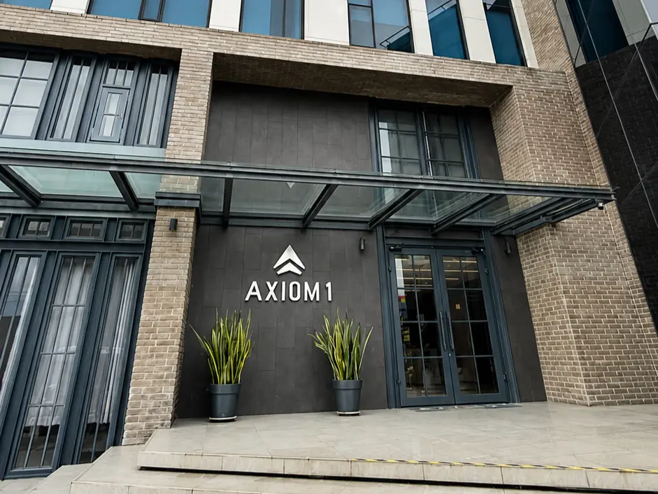 Exterior of the Axiom 1 office building with a glass and brick facade, entrance canopy, and AXIOM1 sign above two potted plants at the top of a staircase