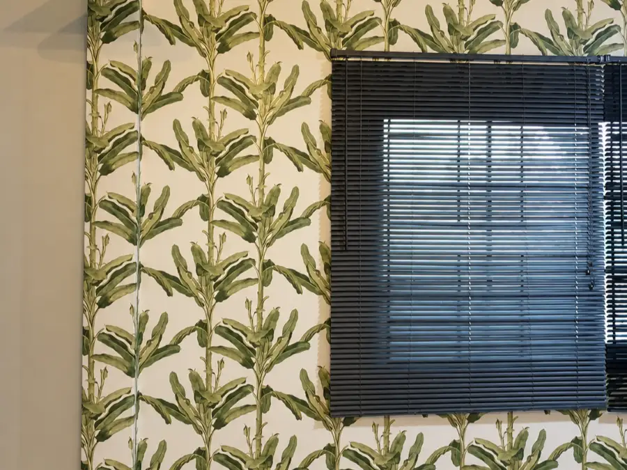 Green banana leaf pattern wallpaper on a wall beside black venetian blinds